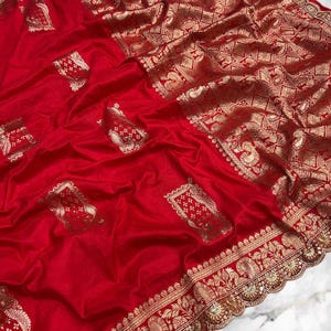 May include: A rich red saree with elaborate gold detailing. The fabric displays detailed floral and geometric patterns along the borders and across the main body, creating a luxurious and ornate appearance. The saree is a traditional garment.