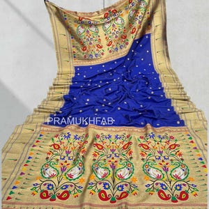 May include: Royal blue sari with intricate gold borders and floral and paisley patterns. The sari features the text "PRAMUKHFAB" and has tassels at the bottom. The design includes colorful flowers, leaves, and bird motifs.