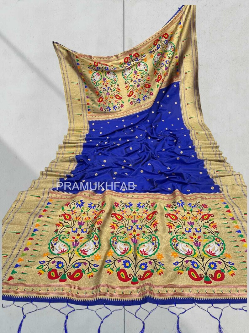 May include: Royal blue sari with intricate gold borders and floral and paisley patterns. The sari features the text "PRAMUKHFAB" and has tassels at the bottom. The design includes colorful flowers, leaves, and bird motifs.