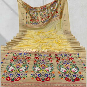 May include: A light yellow saree with intricate embroidery. The saree features a beige border with colorful floral and peacock designs. The text "PRAMUKHFAB" is visible on the saree. The saree is draped, showcasing its texture and design.