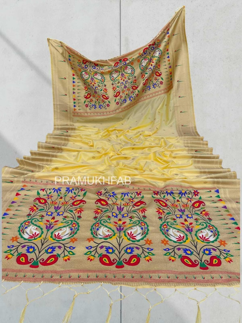 May include: A light yellow saree with intricate embroidery. The saree features a beige border with colorful floral and peacock designs. The text "PRAMUKHFAB" is visible on the saree. The saree is draped, showcasing its texture and design.