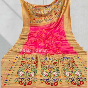 May include: A vibrant pink and gold saree with intricate floral and paisley patterns. The saree features a gold border with colorful accents and the text "PRAMUKHFAB". The design includes a detailed peacock motif.