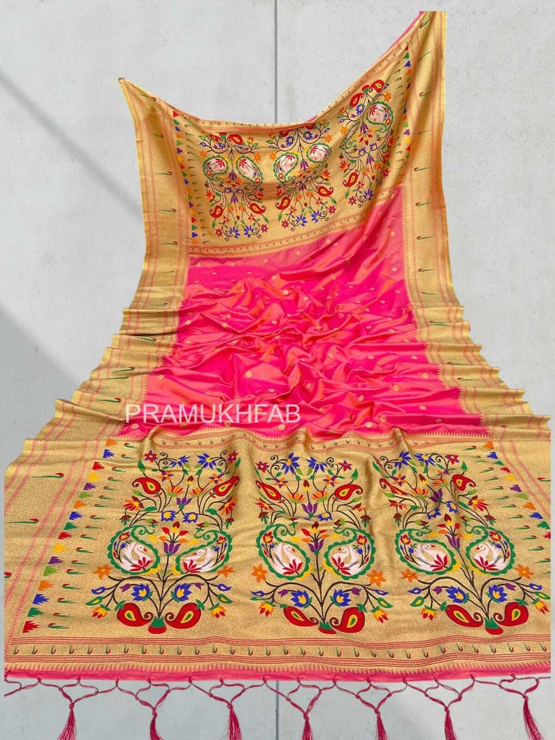 May include: A vibrant pink and gold saree with intricate floral and paisley patterns. The saree features a gold border with colorful accents and the text "PRAMUKHFAB". The design includes a detailed peacock motif.