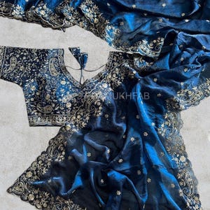May include: A deep blue saree and matching blouse set featuring intricate gold embroidery in paisley and floral patterns. The fabric has a subtle sheen, and the borders are finished with a scalloped edge. The text MUKHFAB is visible.