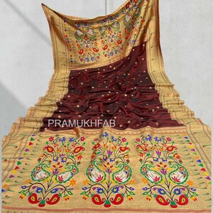 May include: A maroon and gold sari with intricate floral and peacock designs. The sari features a gold border and the text "PRAMUKHFAB". The fabric is draped, showcasing its texture and pattern.