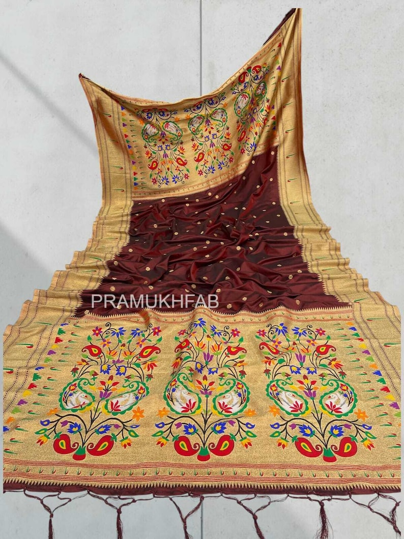 May include: A maroon and gold sari with intricate floral and peacock designs. The sari features a gold border and the text "PRAMUKHFAB". The fabric is draped, showcasing its texture and pattern.