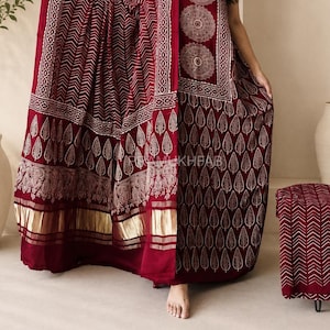 May include: A deep maroon sleeveless dress featuring a white leaf pattern and three circular mandala designs on the bodice. The garment includes a matching shawl with a chevron print and a gold metallic border at the hem, with the text RAMKHEFAB.