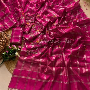 Peut inclure: Un sari fuchsia vibrant orné d'un motif à carreaux argentés métallisés et de broderies florales délicates. Le tissu présente un éclat subtil et inclut une pièce de chemisier assortie. Le texte PRAMUKHFAB est visible sur le tissu à franges.