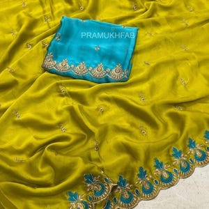 May include: A chartreuse fabric piece featuring scattered gold embroidered motifs and a wide border with blue and gold floral patterns. A folded bright blue fabric square with gold embroidery and the text PRAMUKHFAB is placed on top.