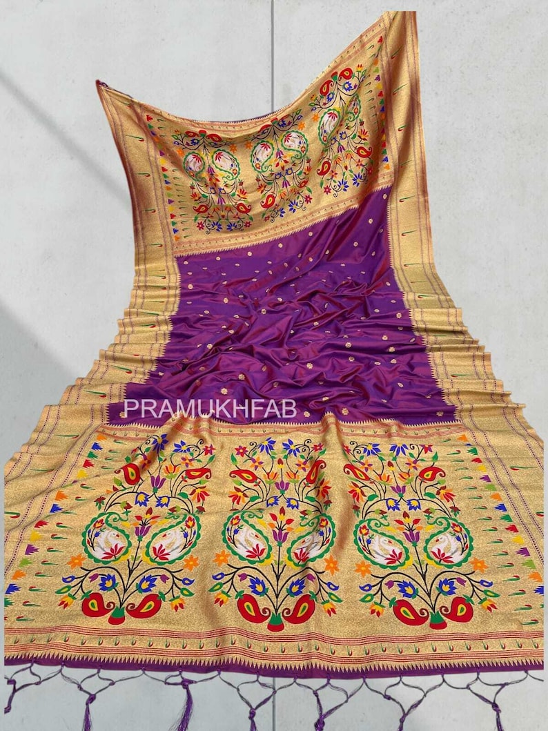 May include: A luxurious purple saree with a gold border and intricate floral and paisley embroidery. The saree features the text "PRAMUKHFAB" and is adorned with colorful accents, creating a vibrant and elegant design.