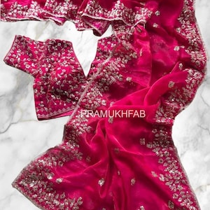 Peut inclure: Un ensemble sari et chemisier fuchsia vif. Le tissu est orné de broderies argentées complexes sur les bordures et sur le chemisier. Le texte "PRAMUKHFAB" est visible sur le tissu.