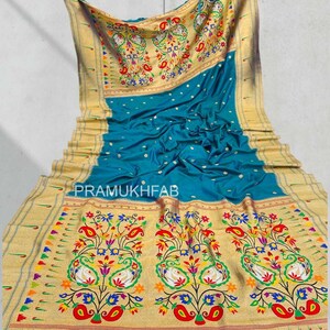 May include: A teal-colored sari with intricate gold borders and floral patterns. The sari features a detailed design with colorful paisley and floral motifs. The text "PRAMUKHFAB" is visible on the fabric.