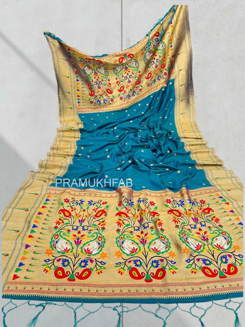 May include: A teal-colored sari with intricate gold borders and floral patterns. The sari features a detailed design with colorful paisley and floral motifs. The text "PRAMUKHFAB" is visible on the fabric.