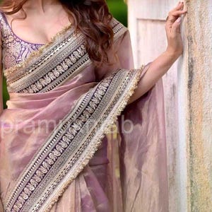 May include: A mauve saree with a sheer, shimmering texture, featuring a wide, dark border adorned with intricate gold embroidery and sequins. The sleeveless blouse is purple with a detailed gold floral pattern and a gold-trimmed neckline.