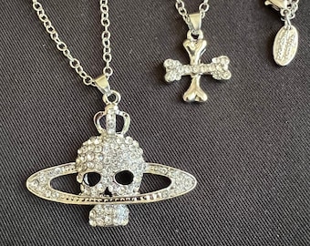 Könnte beinhalten: Zwei silberne Halsketten mit kristallbesetzten Anhängern. Ein Anhänger ist ein Schädel mit einem Ring darum, der andere ist ein Kreuz mit einer Herzform in der Mitte.