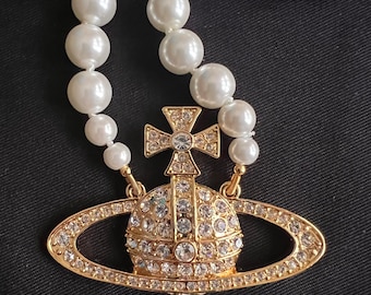Peut inclure: Un collier de perles avec un pendentif doré. Le pendentif présente une croix et un design planétaire, incrusté de cristaux transparents. Le collier est sur fond noir.