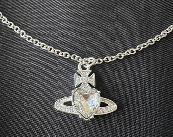 Op de afbeelding: Een zilveren ketting met een kristallen hart hanger. Het hart is omringd door een cirkel van kleinere kristallen. De ketting ligt op een zwarte achtergrond.