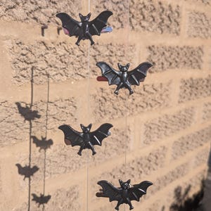 May include: Four black bat-shaped hair clips are attached to a vertical transparent string. Each clip features a detailed bat figure with outstretched wings and pointed ears, secured to a metal hair clip mechanism visible behind the bat shape.