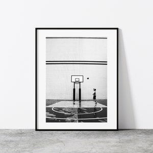 Könnte beinhalten: Schwarzweißfoto von einem jungen Menschen, der auf einem Hallenfeld Basketball spielt. Die Person trägt Shorts und ein T-Shirt und ist im Begriff, den Ball zu werfen. Der Basketballkorb ist im Hintergrund.