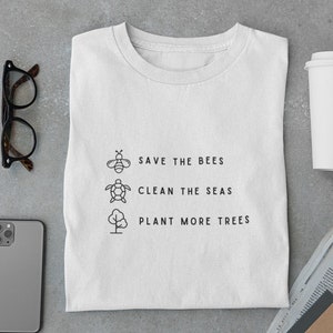 Könnte beinhalten: Weißes T-Shirt mit einem schwarzen grafischen Design, auf dem "Rettet die Bienen", "Saubere Meere" und "Pflanzt mehr Bäume" steht. Das grafische Design beinhaltet eine Biene, eine Schildkröte und einen Baum.