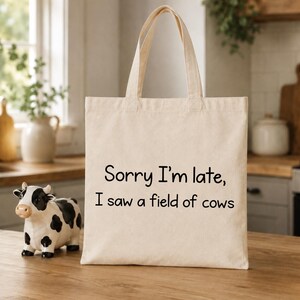 Könnte beinhalten: Eine beige Tragetasche aus Segeltuch mit dem schwarzen Aufdruck Sorry I'm late, I saw a field of cows. Die Tasche hat zwei passende Stoffgriffe und eine rechteckige Form und dient als funktionales Zubehör zum Tragen verschiedener persönlicher Gegenstände.