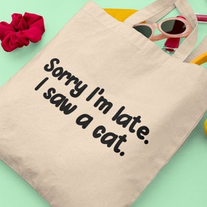 May include: A beige tote bag with black text that reads "Sorry I'm late. I saw a cat." The bag is lying on a light green surface with a glass of iced tea, lemons, sunglasses, hair ties, and headphones.