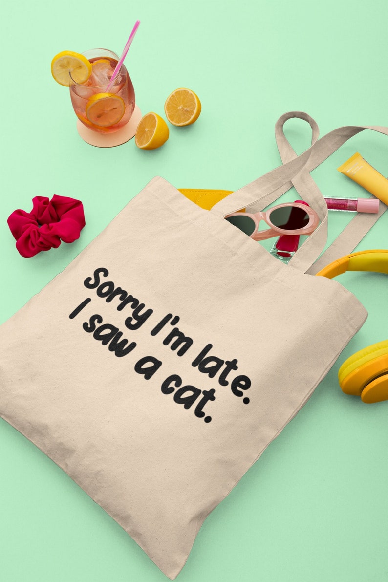 May include: A beige tote bag with black text that reads "Sorry I'm late. I saw a cat." The bag is lying on a light green surface with a glass of iced tea, lemons, sunglasses, hair ties, and headphones.