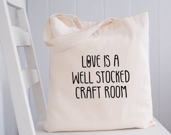 May include: A natural-colored canvas tote bag with two shoulder straps. The front of the bag features black printed text that reads: LOVE IS A WELL STOCKED CRAFT ROOM, with a small heart symbol integrated into the letter O in the word LOVE.