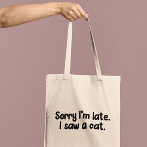 May include: A beige canvas tote bag featuring two shoulder straps and the black printed text Sorry I'm late. I saw a cat. The bag is constructed from a light-colored fabric and displays the text in a bold, rounded, sans-serif font on the front.