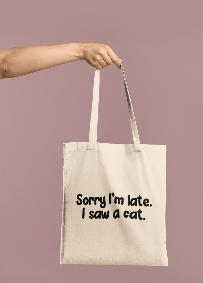 May include: A beige canvas tote bag featuring two shoulder straps and the black printed text Sorry I'm late. I saw a cat. The bag is constructed from a light-colored fabric and displays the text in a bold, rounded, sans-serif font on the front.