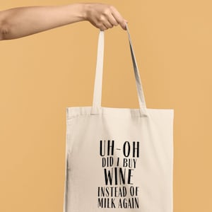 May include: A cream-colored tote bag with black text that reads "UH-OH DID I BUY WINE INSTEAD OF MILK AGAIN." The bag is held by a person against a light orange background. The bag has two handles.