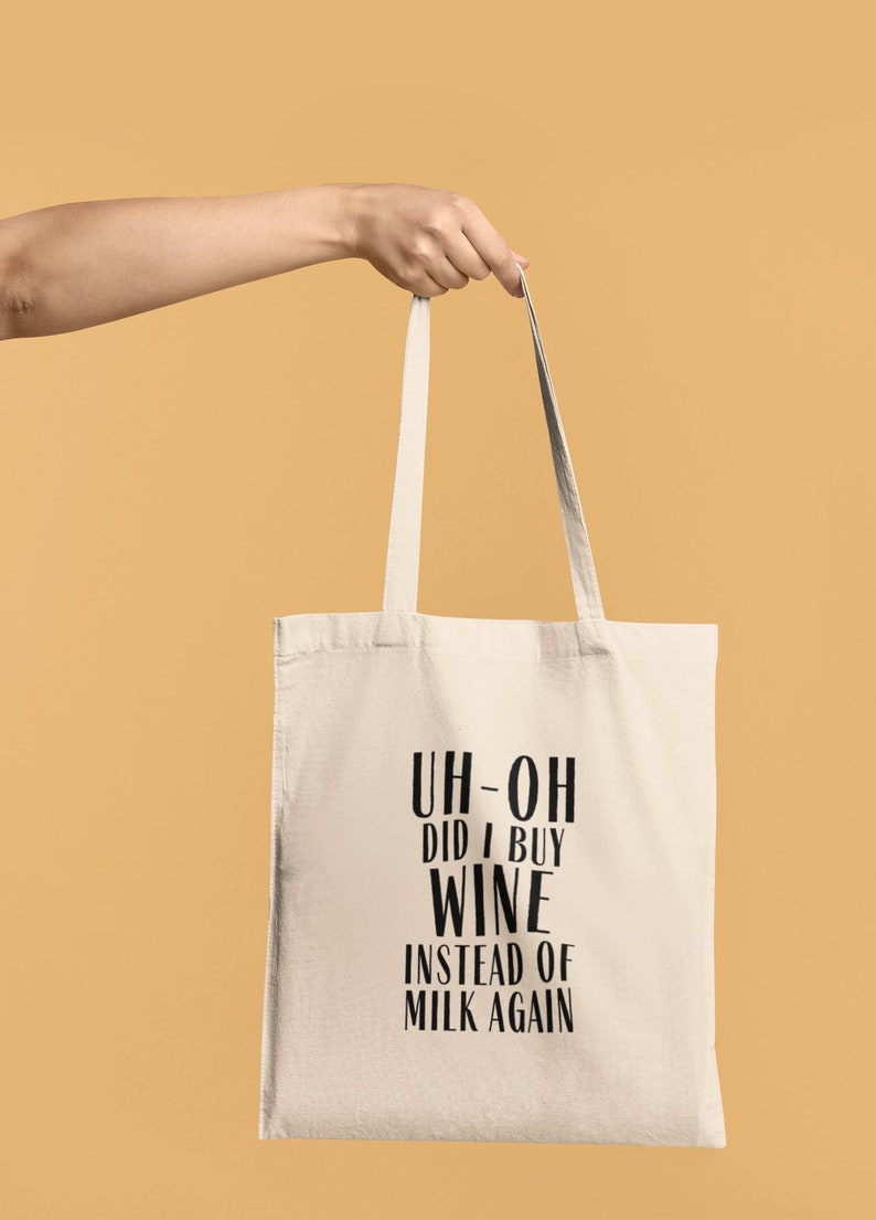 May include: A cream-colored tote bag with black text that reads "UH-OH DID I BUY WINE INSTEAD OF MILK AGAIN." The bag is held by a person against a light orange background. The bag has two handles.