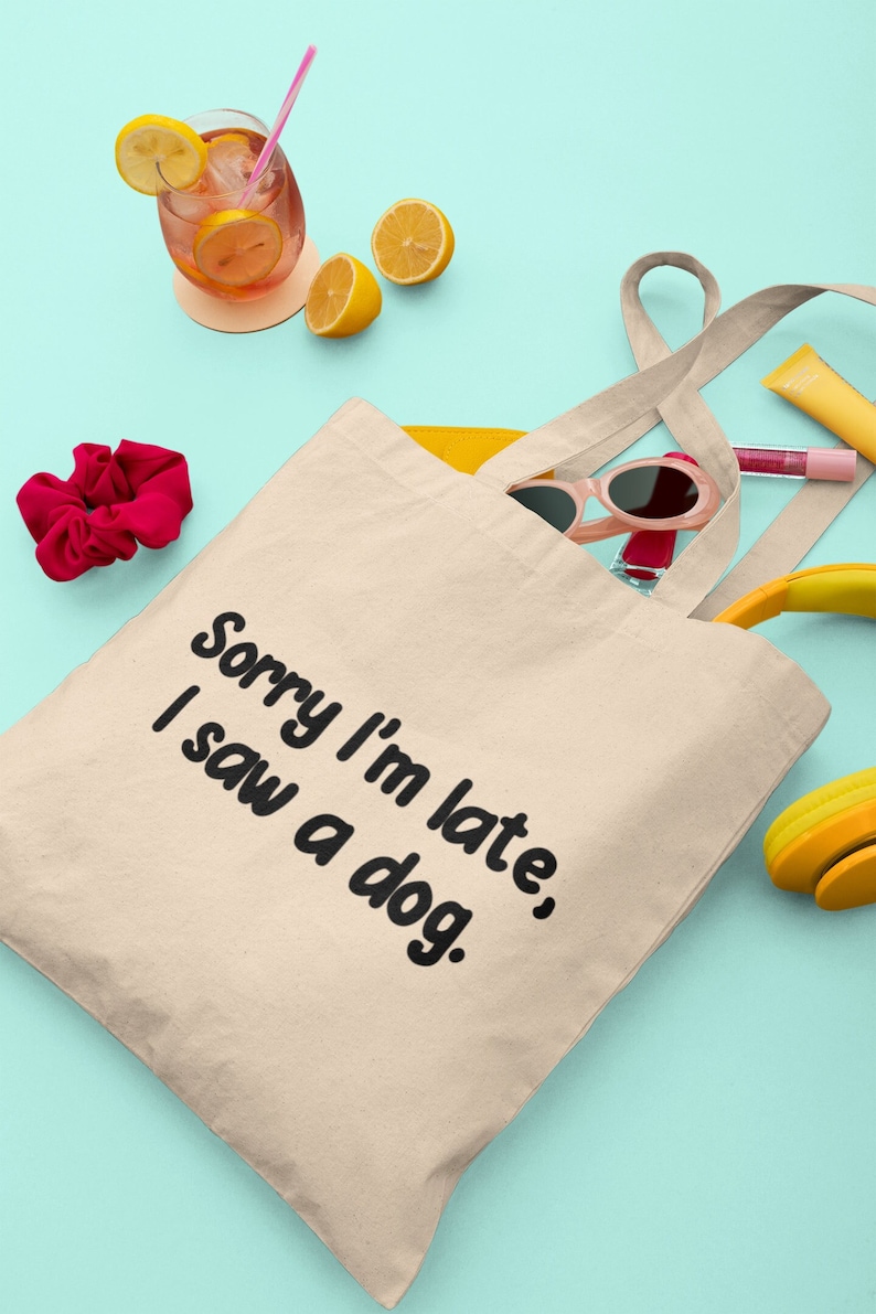May include: A beige canvas tote bag with the text "Sorry I'm late, I saw a dog." printed in black. The bag is surrounded by various items, including lemons, sunglasses, a hair scrunchie, and headphones.