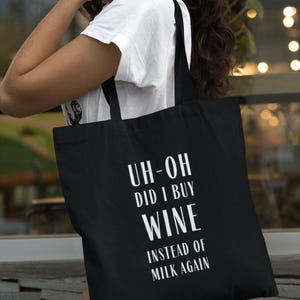 May include: Black tote bag with white text that reads "UH-OH DID I BUY WINE INSTEAD OF MILK AGAIN". The bag is carried over the shoulder. The person is wearing a white t-shirt.