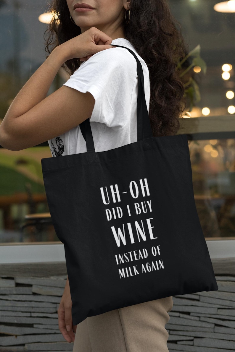 May include: Black tote bag with white text that reads "UH-OH DID I BUY WINE INSTEAD OF MILK AGAIN". The bag is carried over the shoulder. The person is wearing a white t-shirt.