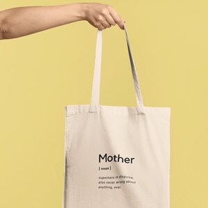 May include: A beige canvas tote bag featuring black printed text that reads: Mother [ noun ] superhero in disguise, also never wrong about anything, ever. The bag has two long shoulder straps and a rectangular shape, presented in a simple design.