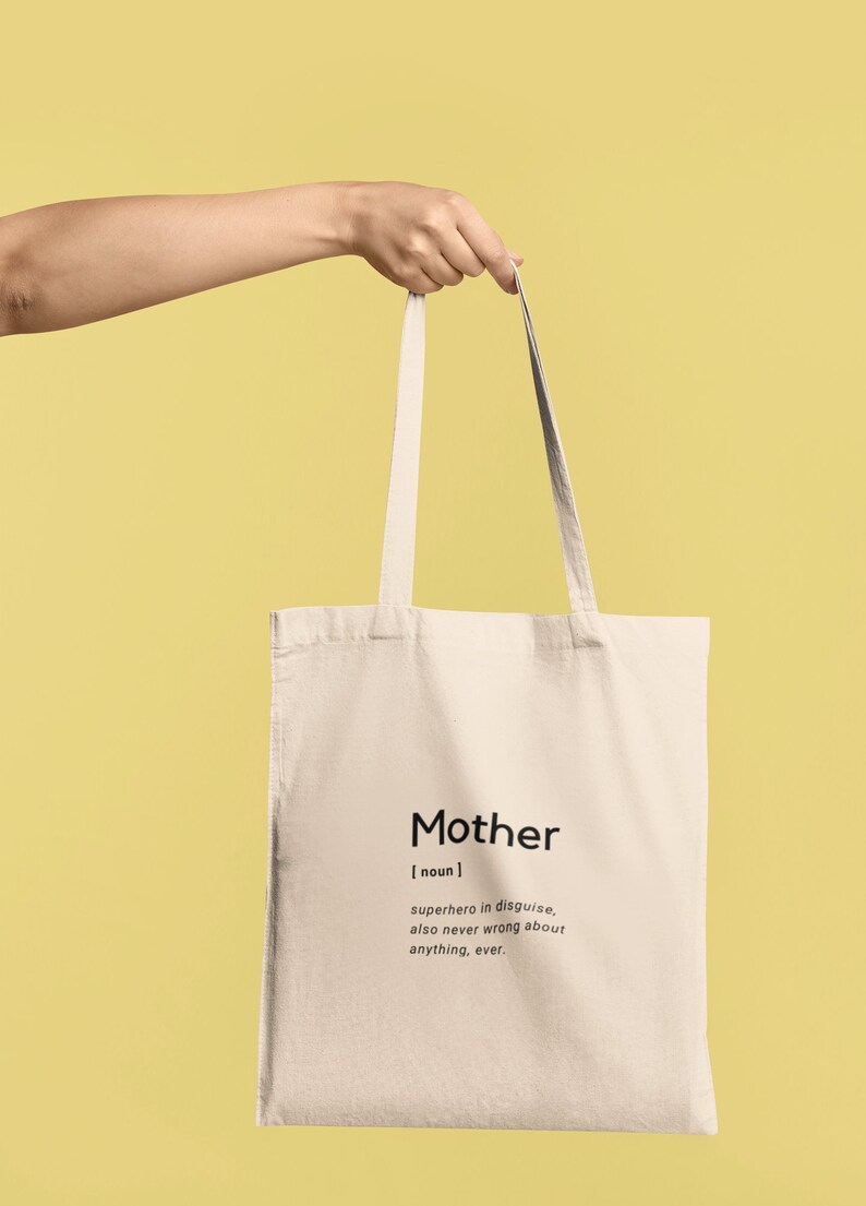 May include: A beige canvas tote bag featuring black printed text that reads: Mother [ noun ] superhero in disguise, also never wrong about anything, ever. The bag has two long shoulder straps and a rectangular shape, presented in a simple design.