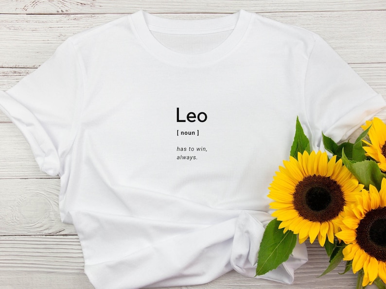 May include: White t-shirt with the word "Leo" printed in black, along with a definition. The shirt is styled with sunflowers. The text reads: "Leo [noun] has to win, always."