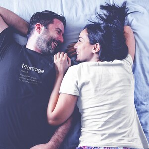May include: A couple lying in bed, smiling at each other. The man is wearing a black t-shirt with the word "Marriage" and a definition printed on it. The woman is wearing a white t-shirt.