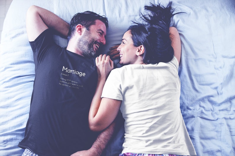 May include: A couple lying in bed, smiling at each other. The man is wearing a black t-shirt with the word "Marriage" and a definition printed on it. The woman is wearing a white t-shirt.