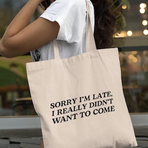 May include: A beige canvas tote bag featuring the black printed text, SORRY I'M LATE, I REALLY DIDN'T WANT TO COME. The bag has two shoulder straps and a rectangular shape, constructed from a light-colored fabric with a simple, functional design.