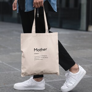 May include: A beige canvas tote bag featuring black printed text that reads Mother [noun] superhero in disguise, also never wrong about anything, ever. The bag has two shoulder straps and a rectangular shape made of a woven fabric material.