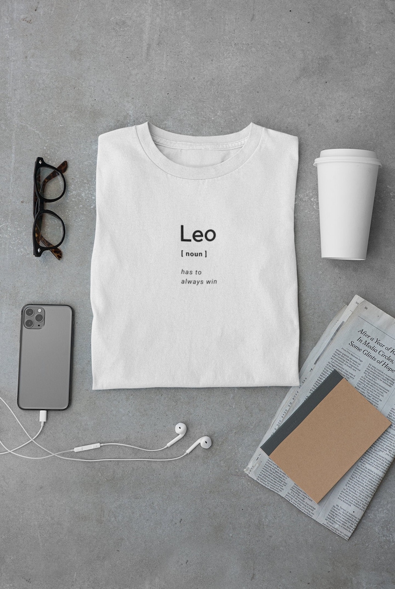 May include: White t-shirt with the word "Leo" and the definition "has to always win" printed in black. The shirt is laid flat on a gray surface, alongside a smartphone, glasses, and a coffee cup.
