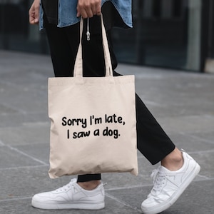 May include: A person is carrying a beige tote bag with the text "Sorry I'm late, I saw a dog." printed in black. The person is wearing a blue denim shirt, black pants, and white sneakers.