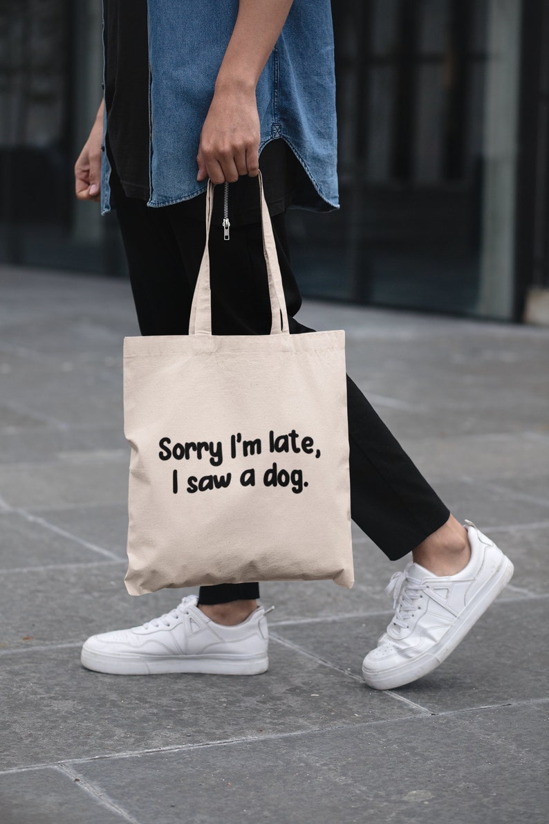 May include: A person is carrying a beige tote bag with the text "Sorry I'm late, I saw a dog." printed in black. The person is wearing a blue denim shirt, black pants, and white sneakers.
