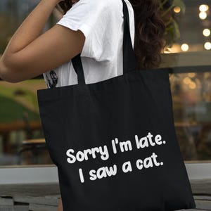 May include: A black tote bag featuring the white text Sorry I'm late. I saw a cat. The bag has two black shoulder straps and a rectangular shape, constructed from a solid fabric material with a clean, simple design for carrying various items.