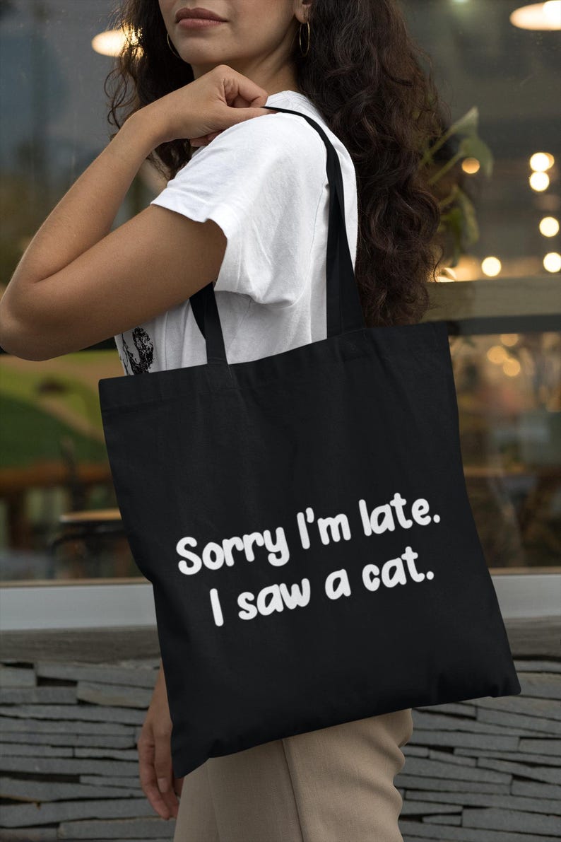 May include: A black tote bag featuring the white text Sorry I'm late. I saw a cat. The bag has two black shoulder straps and a rectangular shape, constructed from a solid fabric material with a clean, simple design for carrying various items.