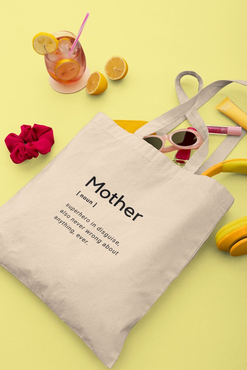 May include: A beige canvas tote bag featuring the black printed text: Mother [noun] superhero in disguise, also never wrong about anything, ever. The bag is surrounded by a pink scrunchie, pink sunglasses, yellow headphones, and a glass of pink lemonade.