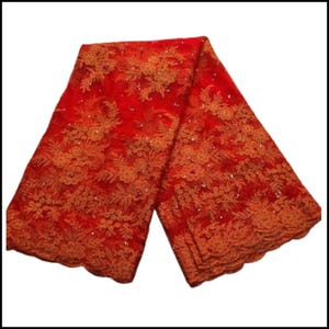 May include: A folded length of red lace fabric, displaying detailed floral patterns and a scalloped edge. The lace is adorned with raised floral elements and small, sparkling embellishments. The fabric's rich red hue and intricate design suggest a luxurious quality, suitable for special occasion wear.