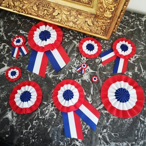 May include: A collection of red, white, and blue rosette ribbons and badges, arranged on a dark, patterned surface. The rosettes vary in size and feature pleated white and red fabric with a blue center. Some have attached blue and white ribbons.